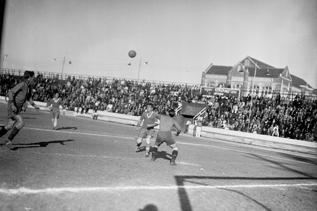 1930s black and white photo of a soccer game being played outdoors with a large crowd in the background. 