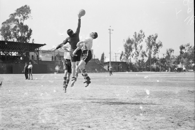 1930s black and white photo of two soccer players clashes midair with a goalie as he hits the ball away from the net.