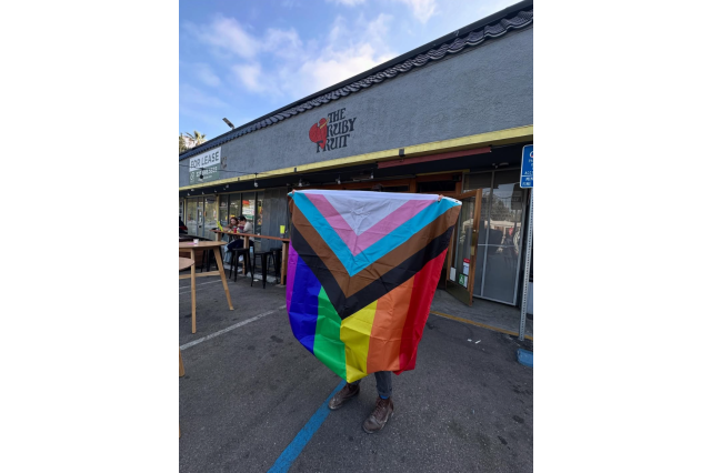 Someone holds an LGBTQ+ Pride Flag in front of themselves, while standing in front of a restaurant named "The Ruby Fruit"