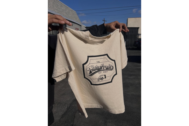 Person holding up a beige souvenir t-shirt that says "The Ruby Fruit, est. 2023".