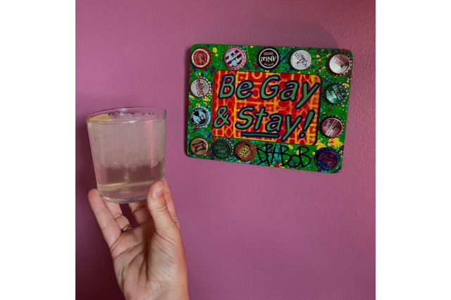 Hand holding up a drink in a high ball glass next to a decorative wall sign that says "Be Gay & Stay".