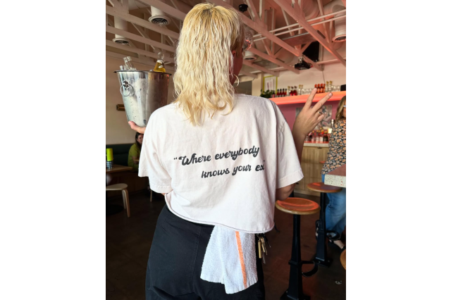 Waitress with long blond pony tail flashing the peace sign, holding an ice bucket with a dish town tucked into her waist. The back of her shirt says "Where everybody knows your ex."