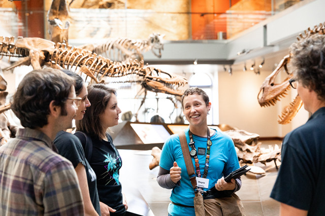 Museum Educator, in front of dinosaur skeletons, facing three guests dinosaur skeletons