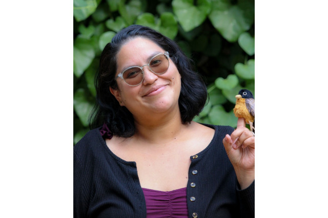 Chantal Ochoa-Clark, wearing sunglasses and holding a stuffed-animal bird on her finder, against a background of green leaves
