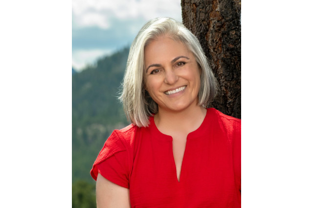 Head and shoulders portrait of Tiffany Wylie wearing a red, short-sleeved blouse, with green hills and a cloudy blue sky behind her