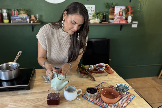 Woman pouring hot tea from a tea pot into a small cup, with the fresh ingredients used to make the tea sitting next to the cup on the counter.