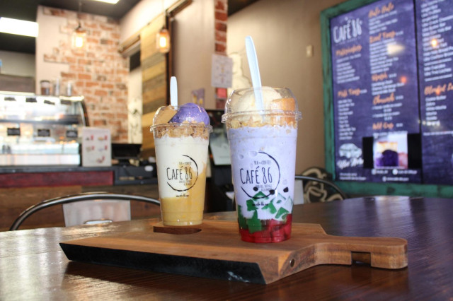 Two colorful dessert drinks with ice cream and fruit jellies in large plastic cups sitting on a wooden cutting board in a cafe.
