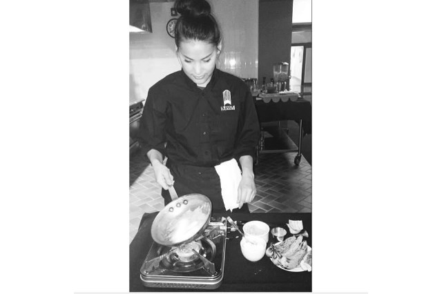 Black and white photo of a young woman cooking over a small stove in a culinary school kitchen.
