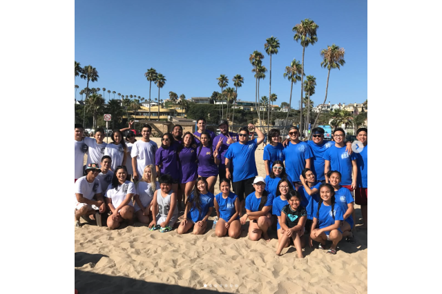 30 young adults in blue and white shirts posing for a group photo on a sandy beach with palm trees in the background.