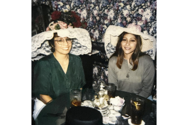 Two women in oversized tea party hats sitting behind a small table with a full tea setting on it.