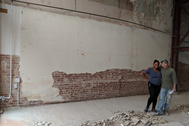 A woman and man stand side by side in front of a bare storefront, exposed brick showing through falling plaster walls. 