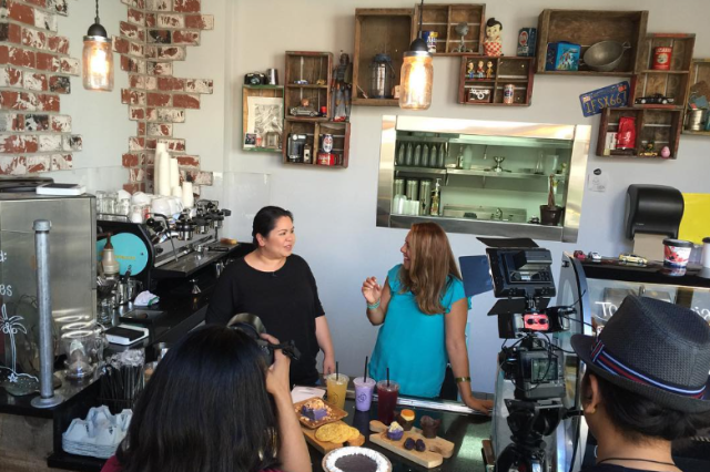 TV crew filming an interview with a chef at the counter of her cafe with drinks and pastries displayed.