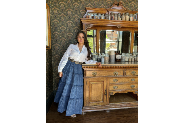 Woman in white shirt and denim skirt standing next to a wooden pantry filled with ingredient jars.