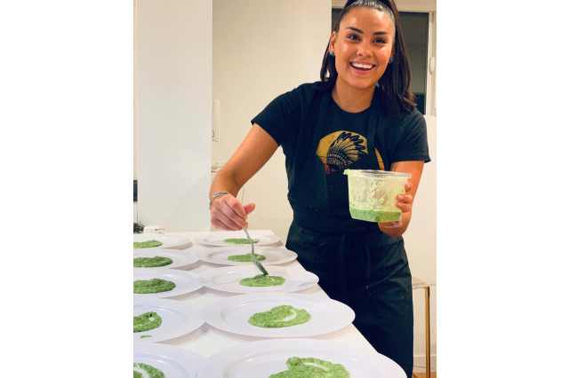 Woman in black shirt and black apron smiling at the camera as she adds green sauce to a table full of plates in preparation for a catering event.
