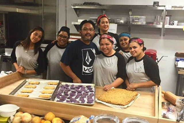 Filipino chefs posing together behind a wooden counter filled with purple ube-flavored pastries.