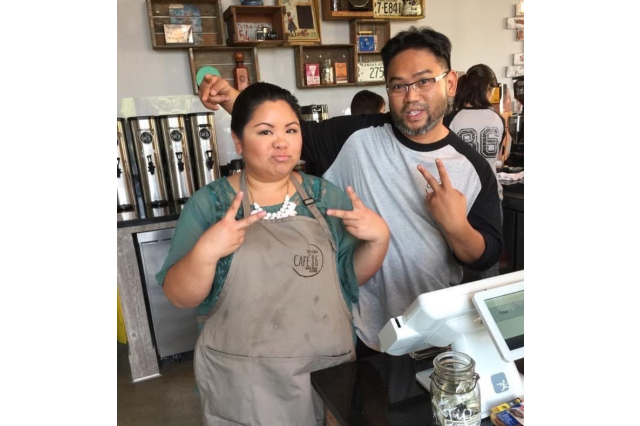 Filipino woman in an apron, standing next to her husband, both giving peace signs in front of a cash register at the opening of their cafe.