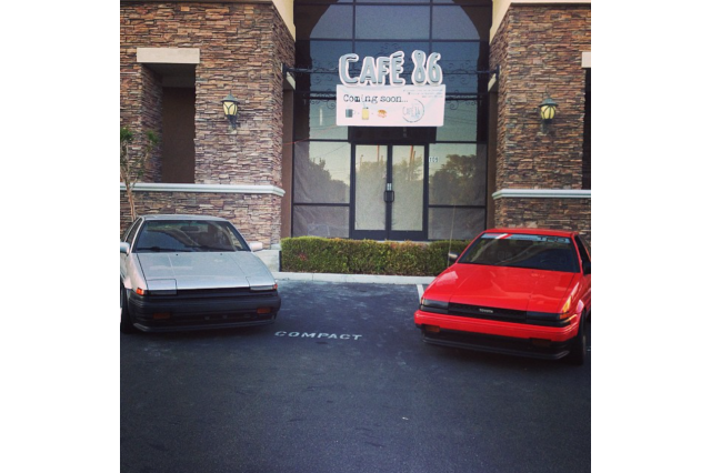 Two 1986 Toyota Corollas, one silver, one red, parked in front of a cafe.