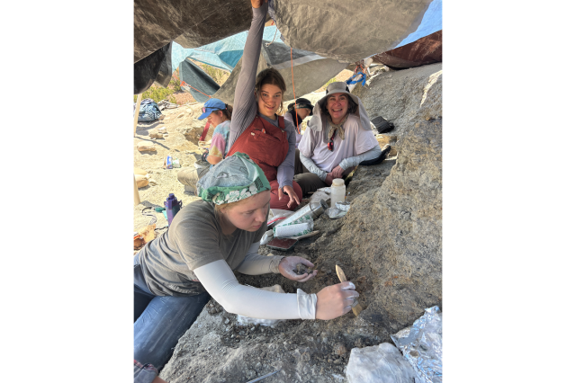 A young woman holds down a tarp, while another woman brushes dirt, and still another smiles at the camera