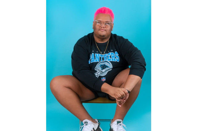 Cody Ziglar, with pink hair, seated with hands on knee against an aqua blue background