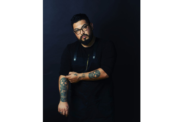 A Mexican American man with facial hair and black glasses poses in front of a dark backdrop.