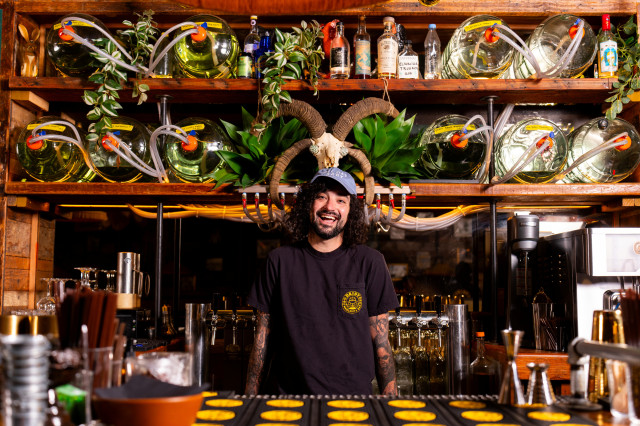 Max Reis, a white man with dark curly hair, stands behind a bar with 5 gallon jars of different drinks and spirits on the wall behind him.