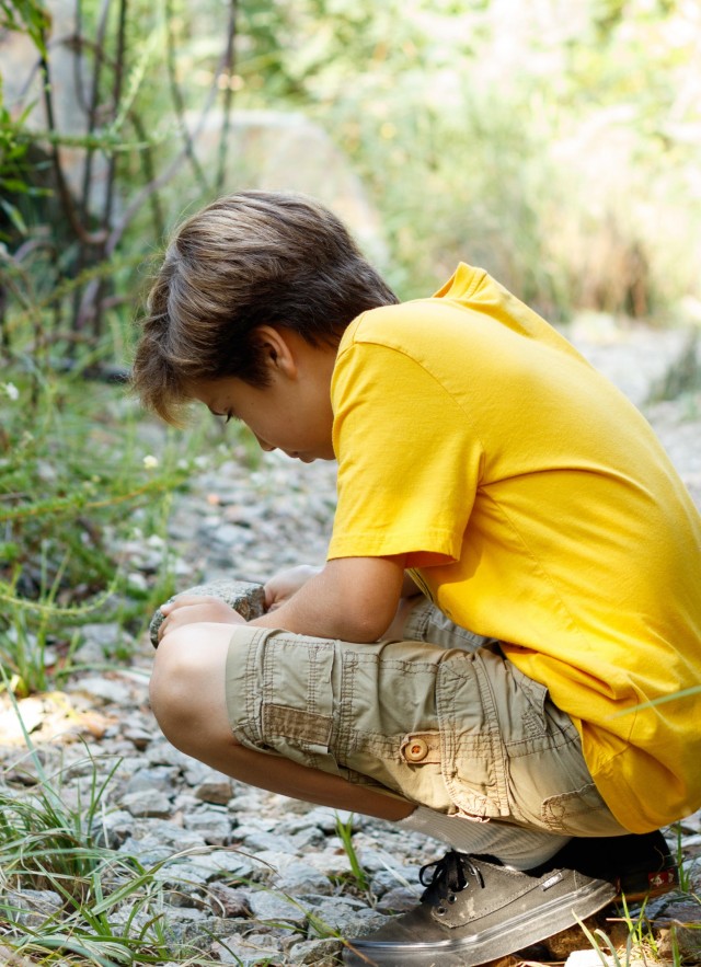 boy with light brown hair and a yellow shirt kneels and looks at something on the ground