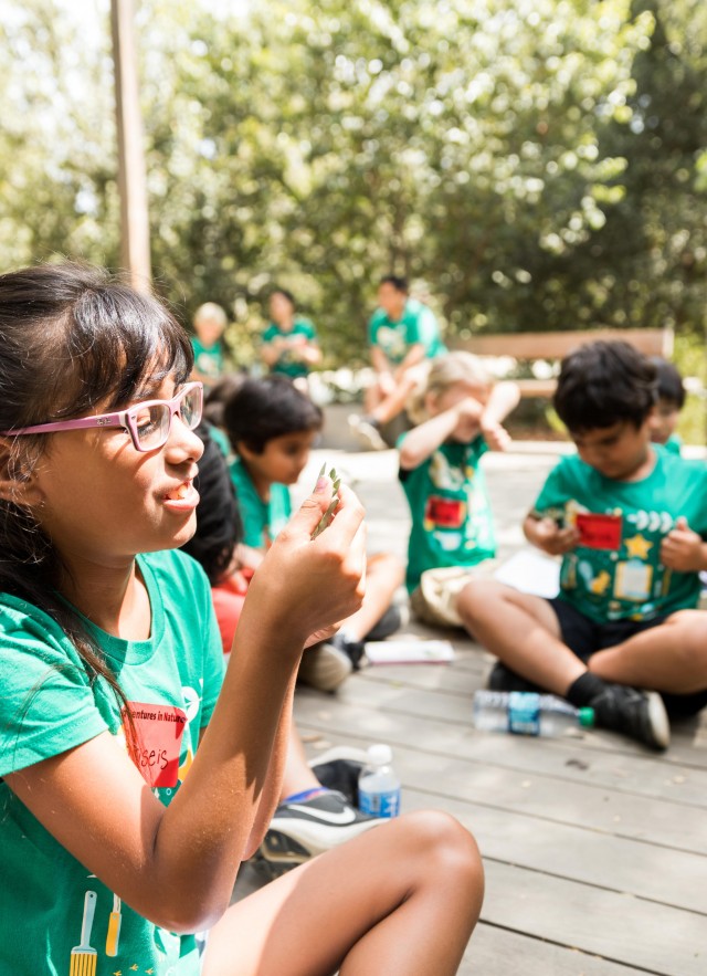 a girl wearign a green shirt and glasses sits outside and looks closely at something in her hand