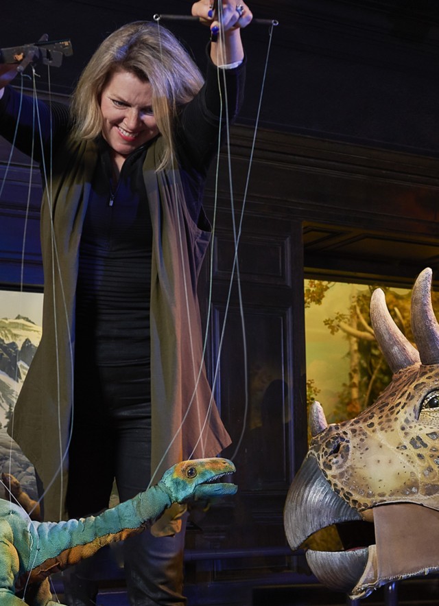 Puppeteer with long-neck dinosaur puppet and triceratops puppet in front of a diorama in the North American Mammal Hall