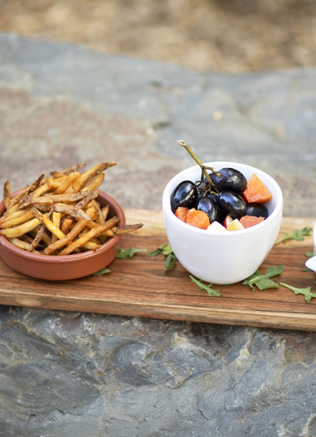 A container of sauce, a bowl of french fries, a bowl of fruit, and a bowl of chicken tenders on a bench