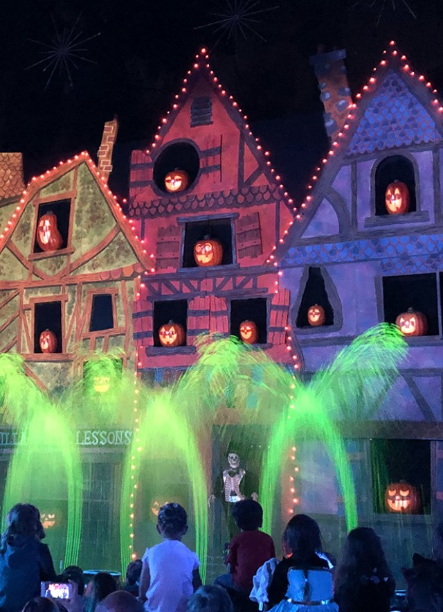 A flat cutout house at night with jack-o-lanterns in the windows, a skeleton character standing in the doorway and audience members watching the performance