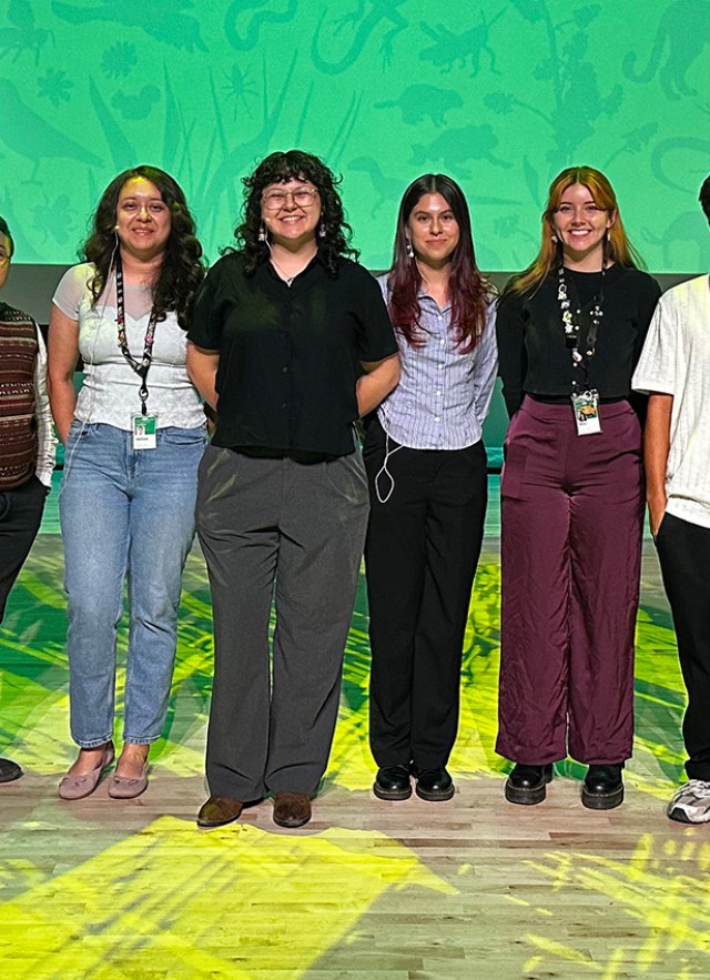 UNLAB participants standing in a row on stage with yellow lighting and a green image on a screen in the background