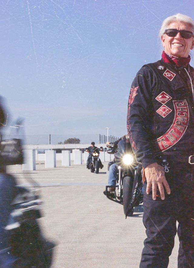 Sam Bearpaw Header with him, a Native an with sunglasses and long white hair pulled back, standing in his Redrum MC vest surrounded by other motorcyclist racing past.