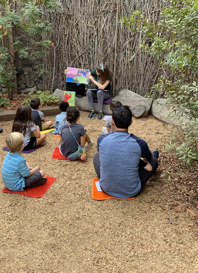 A group of children and adults sitting on individual mats on a wood chip ground, listening to a person read a large picture book during an outdoor story time session. 