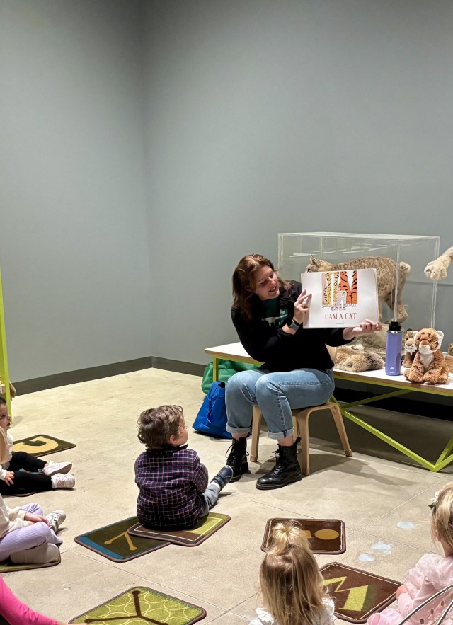 A Museum Educator is reading a book to a group of young children seated on the floor in an educational exhibit space. The room contains display cases with skulls and taxidermied animals, including a large wild cat is catching a bird. Stuffed animal toys are also visible near the reader.