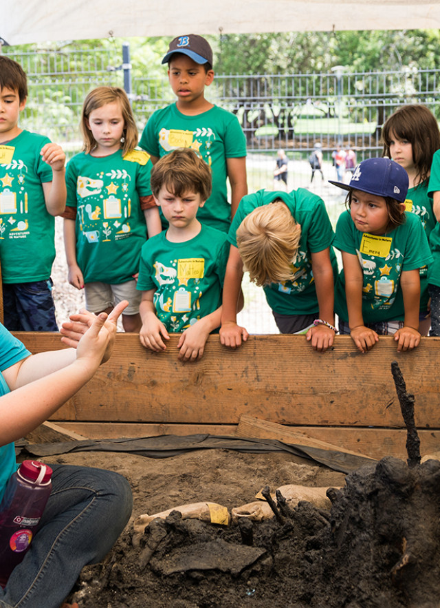 Excavator sitting by a fossil at an excavation site, surrounded by children looking on