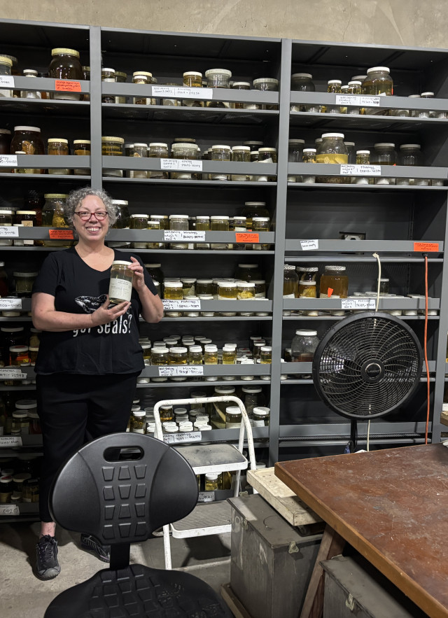 A woman holds a jar in front of a shelf 