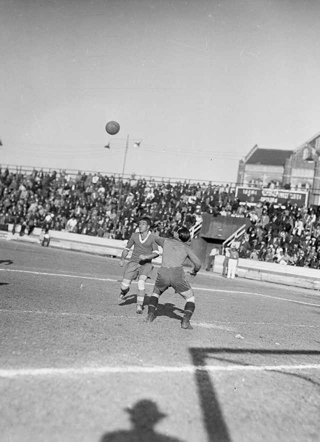 Black and white photo from the 1930s of men playing soccer outside with a packed crowd in bleachers behind them.