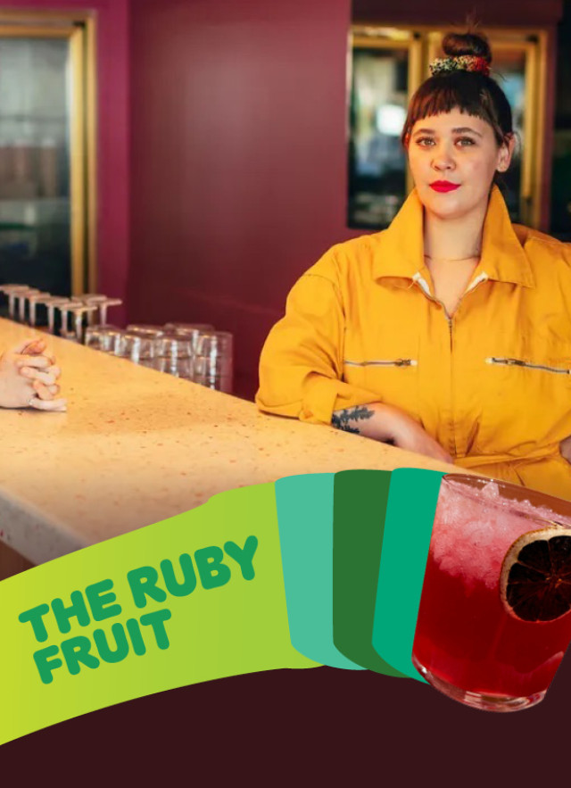 Two people pose behind a long counter inside The Ruby Fruit. One wears a green top and dark pants, while the other wears a bright yellow jumpsuit. In the foreground, a lime green graphic banner features the bar's name next to an image of a red cocktail garnished with a dehydrated citrus wheel.