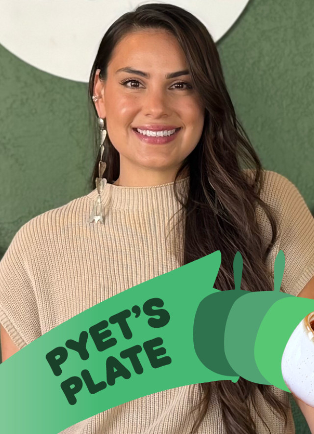 Woman with long, dark hair over one shoulder, standing in front of a green wall with shelves of cups, cookbooks, and plants. 