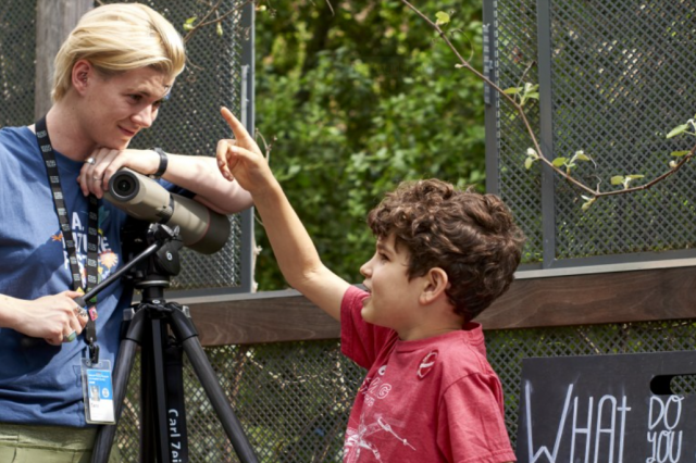 A child points up and exclaims as an educator with a spotting scope listens.
