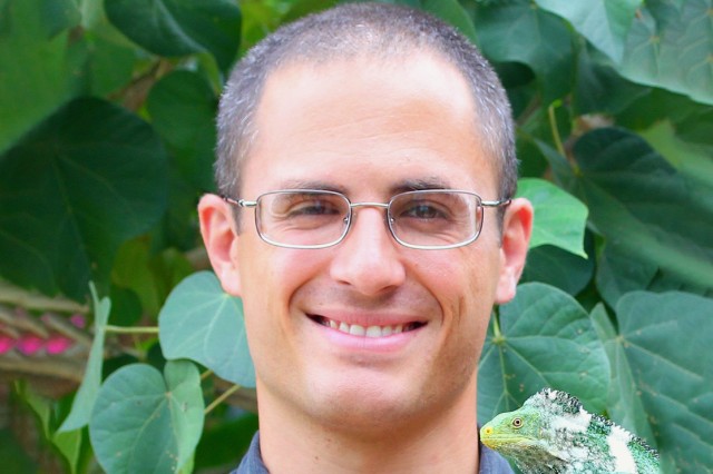 Headshotof Adam Clause, holding a green iguana or lizard and wearing wire-rimmed rectangular shaped glasses with green foliage in the background 