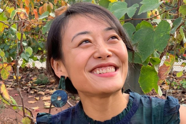 Headshot of Enjie &quot;Jane&quot; Li, wearing a blue and green plaid tank top with ruffle sleeves and blue and green earrings, against a background of green foliage