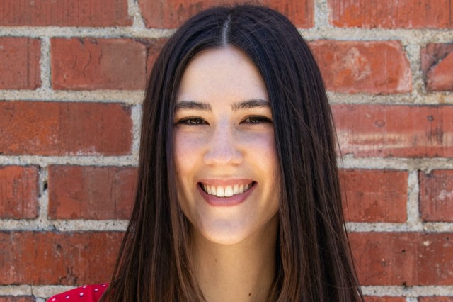Headshot of Jessie Salter with straight long brown hair parted in the middle, against a background of red bricks