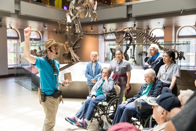 A Museum Educator is in the Dinosaur Hall sharing infomation with several guests.