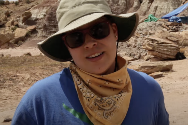 Erika Durazo wearing a hat, sunglasses, and a bandana around their neck with mountains in the background