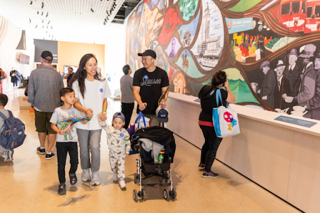 Family of man pushing stroller and woman holding toddler and older child&#039;s hands walking past Carasco mural in NHM Commons