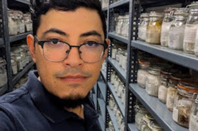 Scientist wearing glasses looks at camera amid jars of specimens on shelves