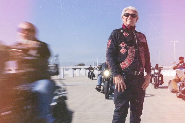 Sam Bearpaw Header with him, a Native an with sunglasses and long white hair pulled back, standing in his Redrum MC vest surrounded by other motorcyclist racing past.