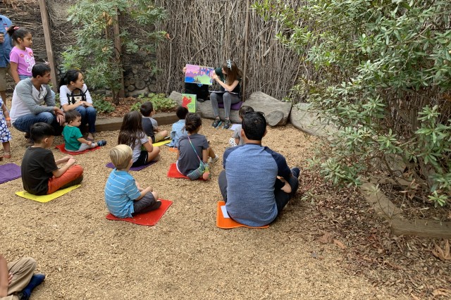 A group of children and adults sitting on individual mats on a wood chip ground, listening to a person read a large picture book during an outdoor story time session. 