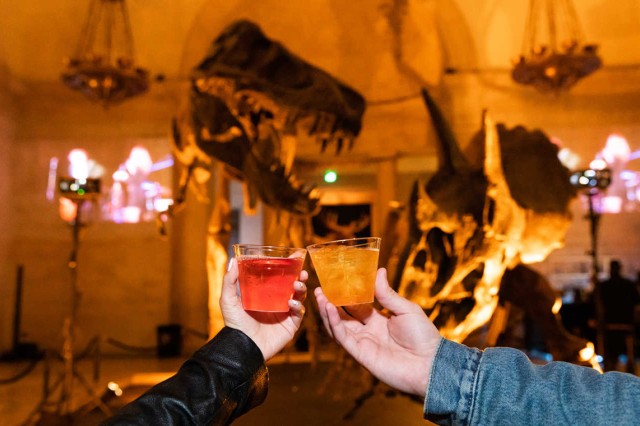 Two women standing at a table with drinks looking at their phone, with a diorama behind them